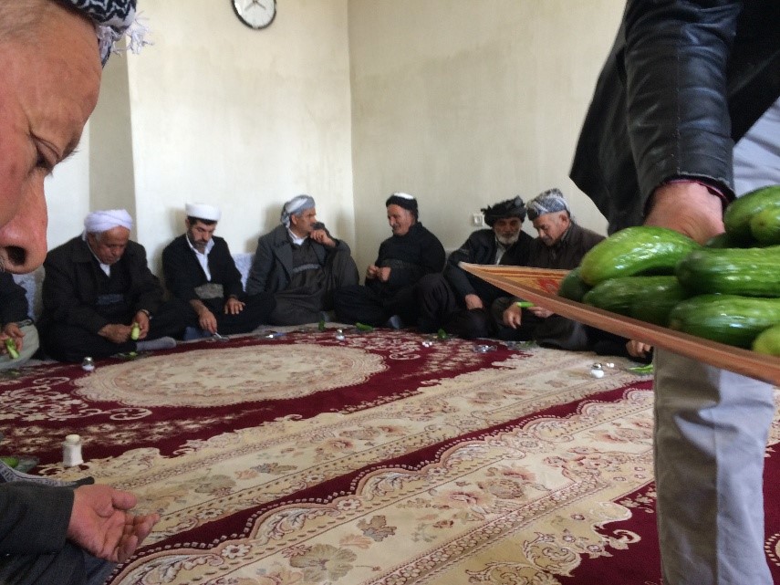People seated around a carpeted room as a tray of cucumbers is carried past.